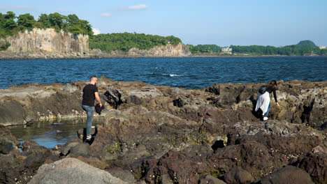 Dos-Personas-No-Identificadas-Caminando-Sobre-Rocas-Volcánicas-Escarpadas-A-Lo-Largo-De-La-Costa-En-La-Isla-Seopseom-Cerca-De-La-Isla-De-Jeju,-Corea-Del-Sur