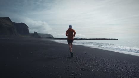 Tiro-De-Seguimiento-De-ángulo-Bajo-De-Un-Hombre-En-Forma-Con-Camisa-Roja-Corriendo-En-La-Playa-Negra-De-Reynisfjara,-Islandia