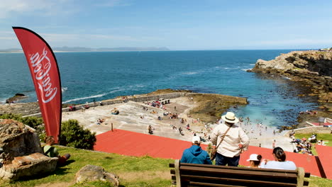 People-on-bench-look-down-at-historic-Old-Harbour-in-Hermanus,-scenic-vista