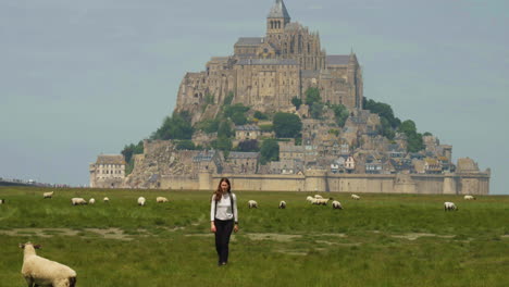 Chica-Turista-Caminando-En-Un-Paisaje-De-Campo-De-Hierba-Con-Ovejas-Pastando-Sobre-Un-Fondo-De-Castillo-Medieval-En-Mont-saint-michel,-Normandía,-Francia