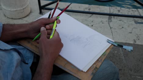 The-Italian-Man-Artist-Hands-With-Pencils-And-Drawing-Pad-Clipped-On-A-Wooden-Board-Sketching-Historical-Monuments-Along-Grand-Canal-In-Venice,-Italy