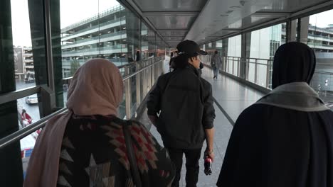 POV-Shot-Behind-Family-Making-Their-Way-Through-To-Terminal-T3-Car-Park-At-Heathrow-Airport,-Crossing-Over-Gangway-Bridge