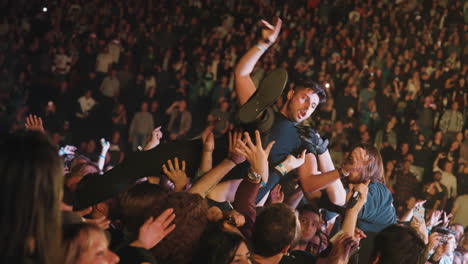 Los-Espectadores-Se-Aglomeran-Animando-Los-Brazos-Levantados-Llevando-A-Un-Hombre-Durante-Un-Concierto-De-Música-En-La-Arena,-La-Intervención-Del-Tipo-De-Seguridad,-Las-Gradas-Y-La-Audiencia-Alrededor-Con-Focos