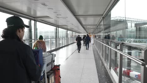 6-November-2022---POV-Beside-Male-Wearing-Baseball-Cap-Pushing-Pushing-Luggage-Trolley-At-Terminal-T3-At-Heathrow-Airport,-Crossing-Over-Gangway-Bridge