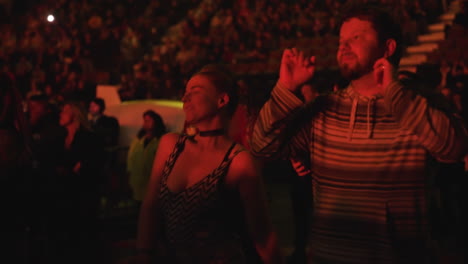 Alegre-Pareja-De-Jóvenes-Espectadores-Fanáticos-Bailando-Durante-Un-Concierto-De-Música,-La-Gente-Se-Aglomera-Y-La-Arena-Se-Encuentra-En-El-Fondo,-Focos-Rojos