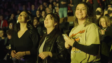 Mujeres-Alegres,-Tres-Jóvenes-Espectadoras-En-Puestos-De-Arena-Durante-Un-Concierto-De-Música,-Cantando-Sonriente-Y-Asintiendo-Con-La-Cabeza,-Multitud-De-Audiencia-Alrededor