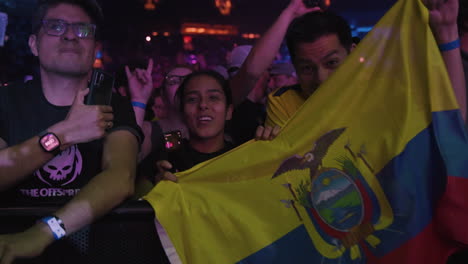 Alegres-Jóvenes-Espectadores-Fanáticos-Cantando-Animando-Sosteniendo-La-Bandera-De-Ecuador-Durante-Un-Concierto-De-Música-En-La-Arena,-Focos-Coloridos-Y-Público-Agolpándose-Alrededor