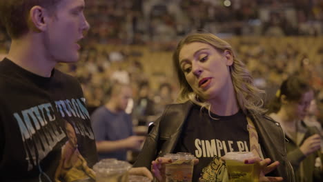 Young-Couple-Spectators-Singing-Dancing-Holding-Beer-Cups-Goblet-During-Music-Concert,-Arena-Stands-and-Audience-Crowd-in-Background