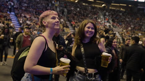 Dos-Jóvenes-Amigas-Espectadores-De-Pie-Bailando-Sosteniendo-Copas-De-Cerveza-Durante-Un-Concierto-De-Música,-Disfrutando-Del-Evento,-Los-Puestos-De-Arena-Y-La-Multitud-Alrededor