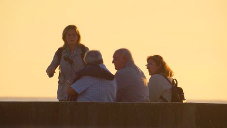 Grupo-De-Viejos-Amigos-Mayores-Sentados-En-La-Playa-Del-Océano-Puesta-De-Sol-Dorada