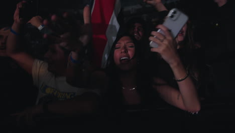 Young-Woman-Lady-Cheering-Singing-and-Dancing-During-Music-Show-with-iPhone-in-Hand,-Indoor-Portrait-with-Spotlights-and-Audience-Crowd-Next-to-Her