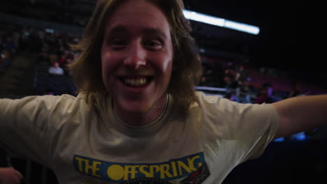 Joven-Alegre-Entrando-En-La-Arena-Antes-Del-Concierto-De-Música-Rock,-Sonriendo-Mirando-A-La-Cámara-Con-Los-Brazos-Extendidos,-Los-Espectadores-Se-Agolpan-En-Las-Gradas-Alrededor