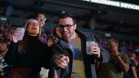 Un-Hombre-Alegre-Con-Una-Novia-Animando-A-Cantar-En-Los-Puestos-De-Arena-Durante-Un-Concierto-De-Música,-Mirando-La-Cámara-Con-Una-Copa-En-La-Mano,-Los-Espectadores-Se-Agolpan-Alrededor