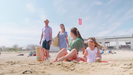 Mother-and-daughter-sitting-on-the-beach-while-mom-welcomes-her-friends-that-just-arrived