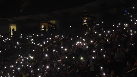 Los-Espectadores-Se-Amontonan-En-Los-Puestos-De-Arena-Con-Teléfonos-Luminosos-Y-Luces-Durante-Un-Concierto-De-Música,-Luces-En-Un-Ambiente-Oscuro-Y-Romántico