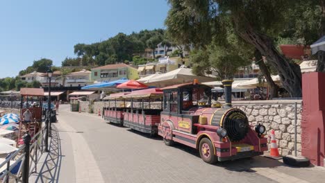 Couple-walking-along-promenade-passing-by-a-touristic-street-train,-Parga,-Greece