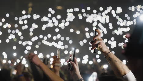 Spectators-Crowd-Hands-Up-Holding-Luminous-Phones-During-Music-Performance-Show,-Arena-Stands-in-Background-with-Bokeh-Defocused-Lights-in-Dark,-Romantic-Atmosphere