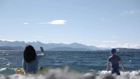 Vista-Detrás-De-La-Madre-Se-Sentó-En-La-Costa-Y-El-Hijo-Tirando-Piedras-En-El-Lago-Nahuel-Huapi-En-Un-Día-Claro-Y-Soleado