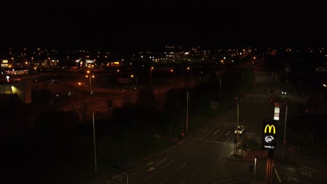 McDonalds-fast-food-drive-through-logo-illuminated-at-night-next-to-Northern-UK-town-highway-rising-aerial-view-right