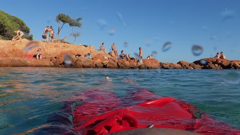 Perspectiva-Personal-En-Cámara-Lenta-Del-Hombre-Vestido-Con-Traje-De-Baño-Rojo-Con-Piernas-Y-Pies-Relajándose-Mientras-Flota-En-Aguas-Tranquilas-Con-Rocas-De-Palombaggia-Y-Gente-En-El-Fondo