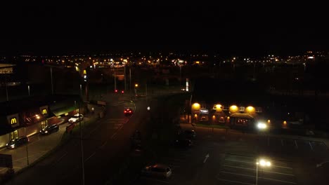 Volando-Sobre-La-Unidad-De-Comida-Rápida-Mcdonalds-Iluminada-Por-La-Noche-Junto-A-La-Vista-Aérea-De-La-Autopista-De-La-Ciudad-Del-Reino-Unido-Que-Sube-A-La-Izquierda