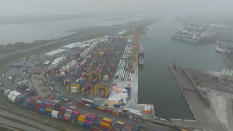 Vista-Aérea-De-La-Terminal-De-Contenedores-De-La-Zona-Portuaria,-Contenedores-Metálicos-Con-Carga-Y-Barcos-Amarrados-En-Los-Muelles-Temprano-Y-Oscuro-Por-La-Mañana-En-El-Puerto-De-Klaipeda