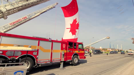 Camiones-De-La-Ceremonia-Conmemorativa-De-Los-Bomberos-De-Toronto-Que-Cuelgan-Enormes-Banderas-Canadienses-Con-La-Sombra-De-Los-Aviones-Que-Pasa-Por-Encima