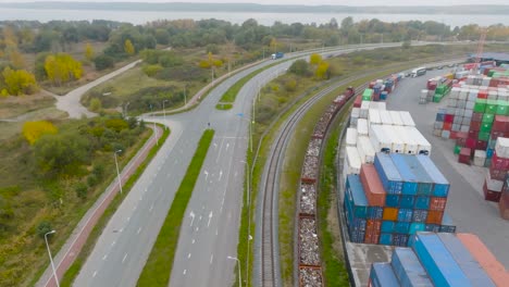 Vista-Aérea:-Un-Tren-Tira-De-Vagones-De-Carga-Paralelos-A-Una-Carretera-De-Automóviles-Y-Una-Terminal-De-Carga-En-La-Ciudad-De-Klaipeda