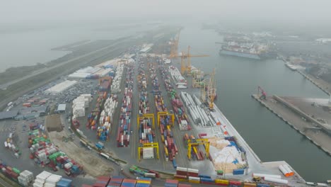 Vista-Aérea-De-La-Terminal-De-Contenedores-De-La-Zona-Portuaria,-Contenedores-Metálicos-Con-Carga-Y-Barcos-Amarrados-En-Los-Muelles-Temprano-Y-Oscuro-Por-La-Mañana-En-El-Puerto-De-Klaipeda