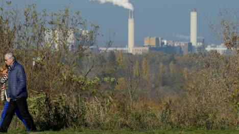 Senior-couple-walking-on-sunny-park-path-with-contrasting-polluting-factory-on-skyline