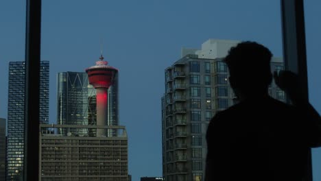 Young-man-looking-out-window-with-a-city-skyline-in-the-background,-during-the-dusk-hour