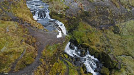 Pareja-Caucásica-Recién-Casada-Abrazándose-Y-Parada-En-Un-Acantilado-Junto-A-Una-Hermosa-Cascada-En-El-Paisaje-De-Islandia