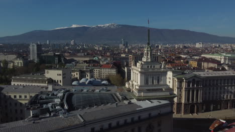 Drohnenaufnahme-Von-Sofia-In-Bulgarien---Drohne-Fliegt-Um-Das-Largo-Gebäude-Herum-Und-Blickt-Auf-Die-Berge