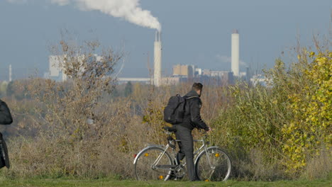 Joven-Se-Sube-A-La-Bicicleta-Con-Humos-De-La-Industria-En-El-Fondo,-Transporte-Sostenible