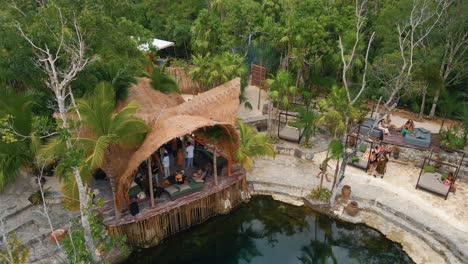 Vista-Aérea-De-Un-Edificio-De-Madera-Tropical-Junto-A-Un-Cenote,-Tulum,-México,-Rodeado-De-Palmeras,-Con-Gente-Relajándose