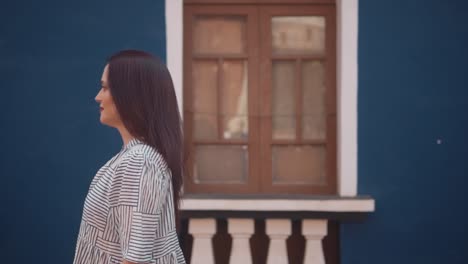 Brunette-Indian-woman-walking-in-front-of-a-blue-building,-with-a-smile,-in-the-historical-streets-of-Fontainhas,-India