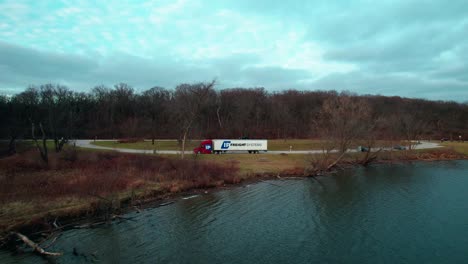 Ziehen-Sie-Die-Antenne-Eines-Roten-Sattelschleppers-Und-Eines-Weißen-Anhängers-Weg,-Die-An-Der-Seite-Eines-Sees-Geparkt-Sind