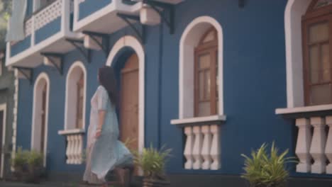 Brunette-Indian-woman-happily-walking-away-and-spinning-around,-in-front-of-a-blue-building-in-the-historical-streets-of-Fontainhas,-India