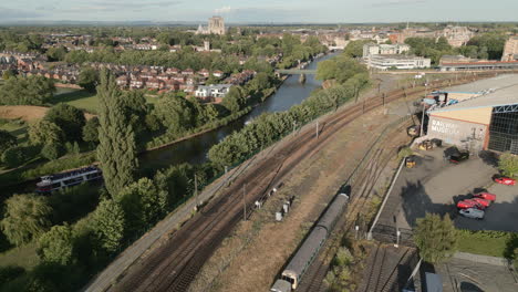 Un-Dron-Aéreo-Disparó-A-Lo-Largo-De-La-Línea-Ferroviaria-De-York-Y-La-Casa-Fluvial-Con-El-Transbordador-Turístico-Pasando-Y-La-Catedral-De-La-Catedral-De-York-En-Segundo-Plano-En-La-Soleada-Puesta-De-Sol-Del-Norte-De-Yorkshire,-Reino-Unido