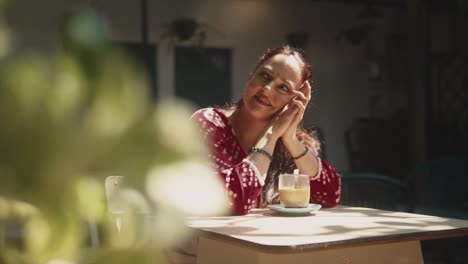 Beautiful-Indian-woman-with-a-dreamy-smile-leaning-her-head-on-her-hands,-with-her-morning-coffee-on-a-outdoor-table,-in-Fontainhas,-India