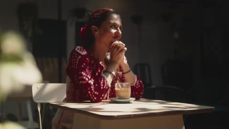 Beautiful-Indian-woman-looking-away-with-a-dreamy-smile,-with-her-morning-coffee-on-a-outdoor-table,-wearing-a-red-summer-dress,-in-Fontainhas,-India