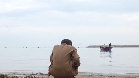 Vista-Desde-Detrás-De-Un-Joven-Pakistaní-Sentado-En-Una-Posición-En-Cuclillas-Mirando-Al-Mar-Desde-La-Playa-De-Gwadar