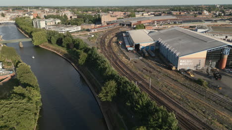 Luftdrohnenaufnahme-Des-Nationalen-Eisenbahnmuseums-Und-Des-Flusses-Ouse-Mit-Einem-Boot,-Das-Den-Fluss-Hinunter-Fährt,-Mit-Dem-Bahnhof-Von-York-Im-Blick-Auf-Einen-Sonnigen-Abend---York-City,-North-Yorkshire,-Vereinigtes-Königreich