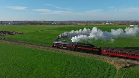 Un-Dron-Paralelo-A-La-Vista-Trasera-De-Un-Tren-De-Pasajeros-De-Vapor-Que-Sopla-Mucho-Humo-Y-Vapor-En-Un-Día-Soleado-De-Otoño