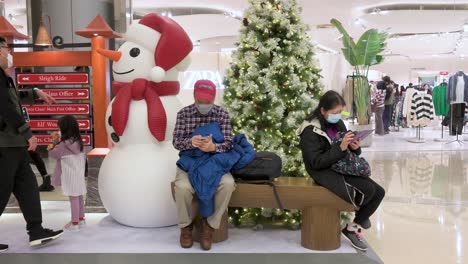 Los-Compradores-Chinos-Se-Sientan-En-Un-Banco-Junto-A-Un-árbol-De-Navidad-Y-La-Instalación-De-Un-Muñeco-De-Nieve-En-Un-Centro-Comercial-Mientras-Otros-Clientes-Pasan-Junto-A-Ellos-En-Hong-Kong