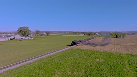 Eine-Luftaufnahme-Einer-Einzelnen-Eisenbahnstrecke,-Die-Durch-Ländliches-Ackerland-Führt,-Während-Sich-Ein-Dampfzug-Nähert-Und-Rauch-Bläst