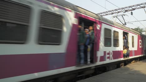 Crowded-local-train-crosses-the-railway-station-platform-in-speed-with-people-standing-at-door-during-rush-hour-in-hot-summer-day