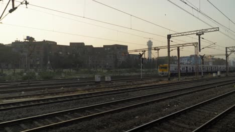 Vista-Lateral-Del-Patio-Del-Ferrocarril-Desde-Un-Tren-En-Movimiento-Que-Sale-Lentamente-Del-Cruce,-Tren-Local-Parado-Y-Esperando-La-Señal-En-Otra-Línea-Con-Viejos-Edificios-Rústicos-Al-Fondo