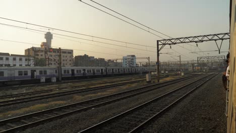 Vista-Lateral-Del-Patio-Del-Ferrocarril-Desde-Un-Tren-En-Movimiento-Que-Sale-Lentamente-Del-Cruce,-Tren-Local-Parado-Y-Esperando-La-Señal-En-Otra-Línea-Con-Viejos-Edificios-Rústicos-Al-Fondo