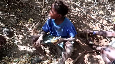 Feliz-Niño-De-La-Tribu-Hadzabe-Pelando-Una-Raíz-De-Baobab-Con-Un-Cuchillo-Mientras-Un-Hombre-Intenta-Encontrar-Más